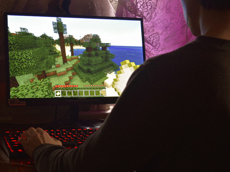 En computerskærm viser en scene fra Minecraft, men man ser skulderen og ryggen af en person, men hånden på et gamer-tastatur